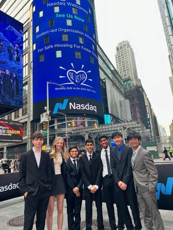 See Us Now members in front of a blue Nasdaq screen.